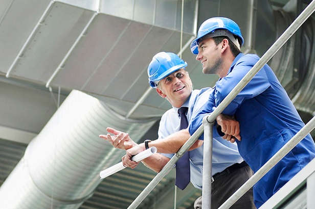 two men wearing blue hardhats leaning on railing in plant and talking