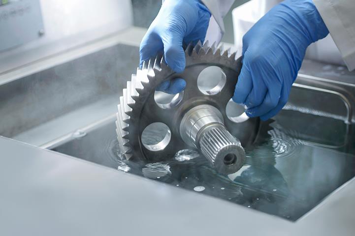 worker wearing blue gloves holding gear in cleaning liquid